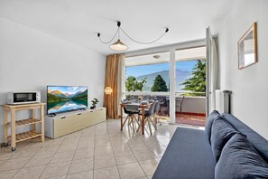 A modern living room with a flat-screen TV, a sofa, a dining area, and a balcony with a mountain view. - Lagotissimo 17 - Apartment with Balcony View and Pool, Riva del Garda, Italy (Riva del Garda)