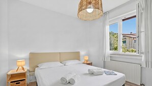 Ein ordentlich bezogenes Bett mit weißen Laken, zwei Nachttische mit Lampen, ein Lampenschirm aus Weide und ein Fenster mit Vorhängen.
