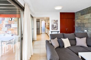 A modern living room with a grey sofa, a dining area with a white table and chairs, and a kitchen in the background. - GuestReady - Villa Salobre Par4 Private Pool (San Bartolomé de Tirajana)