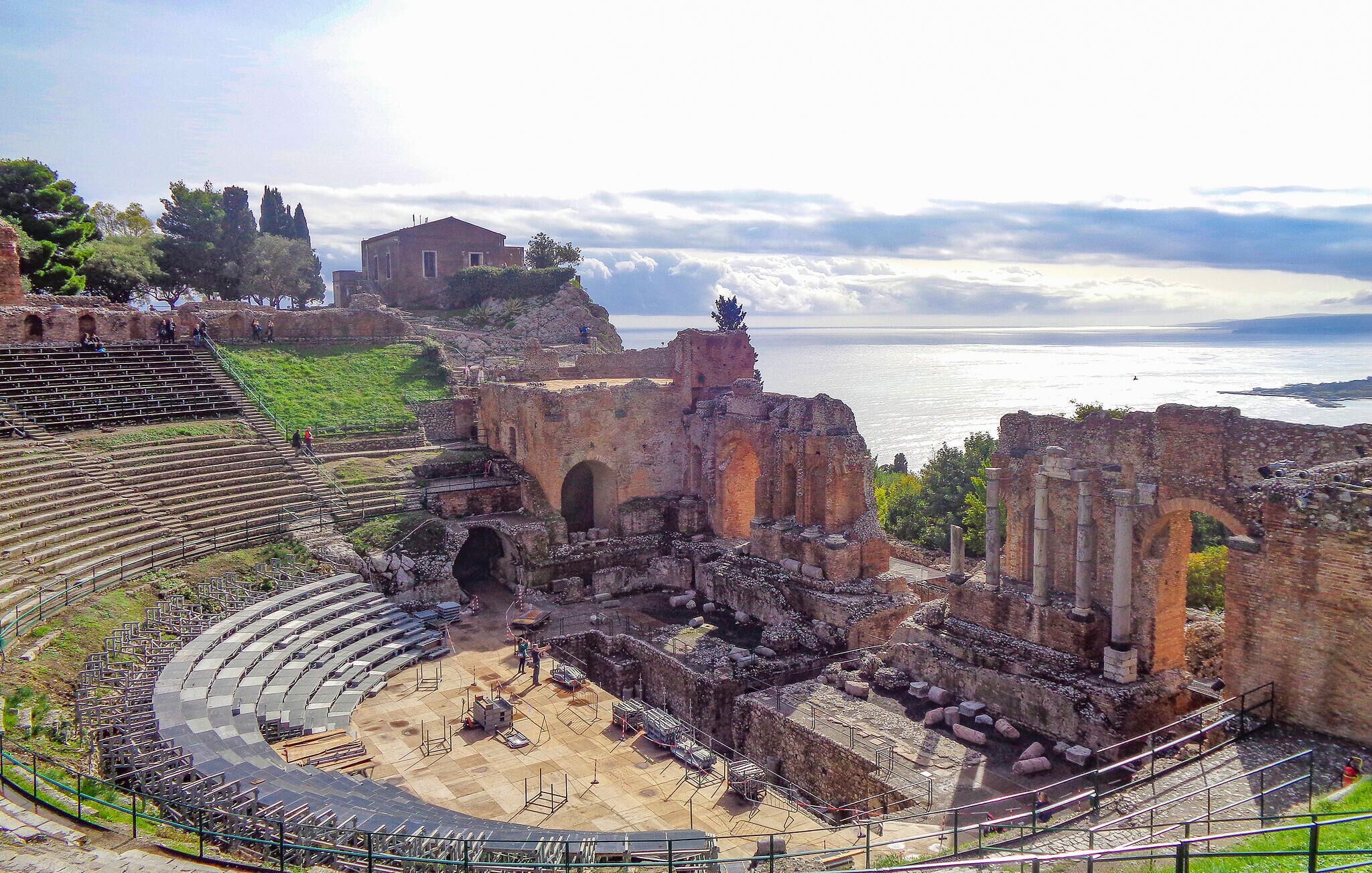 Un anfiteatro antiguo con un escenario y zona de asientos parcialmente conservados, rodeado de ruinas y con vistas a un cuerpo de agua.