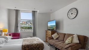 Quarto de hotel com cama, sofá, televisão, janela com cortinas e relógio de parede.