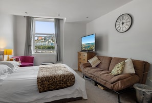 A hotel room with a bed, a sofa, a television, a window with curtains, and a wall clock. - Harbour View at No.11, an exceptional coastal holiday home in Brixham (Brixham)