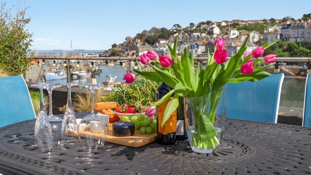 Uma mesa com vista para um porto, uma garrafa de vinho, um vaso de flores e uma tábua de queijos com frutas e pão.