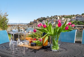 Een tafel met uitzicht op een haven, een fles wijn, een vaas bloemen en een kaasplank met fruit en brood.
