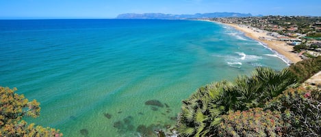 A coastal landscape with clear turquoise waters, a sandy beach, and a view of a town in the distance.