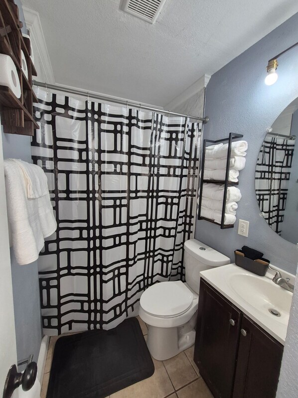 Un baño con cortina de ducha de estampado blanco y negro, inodoro blanco, lavabo con espejo y toallas en un perchero.