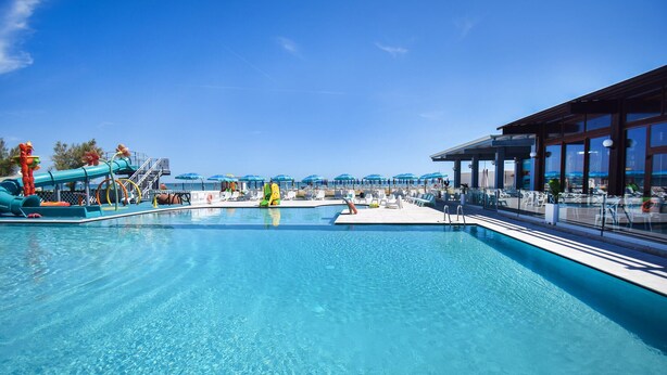 Une piscine d’un bleu éclatant, avec un toboggan et des chaises longues, un espace abrité avec des parasols, et une vue sur l’océan.