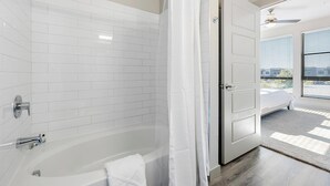 A modern bathroom with a bathtub, white tiled walls, and a door leading to a bedroom with a view. - Landing Furnished Apartments - Northwest Austin (Austin)