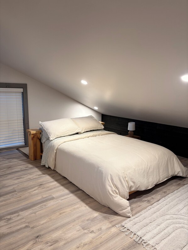 Ein Schlafzimmer mit Bett, Nachttisch, Lampe und einem Fenster mit Jalousien.