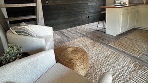 Un espace de vie de style loft avec des poutres apparentes, une cuisine intégrée et un coin salon confortable avec un canapé et un pouf.