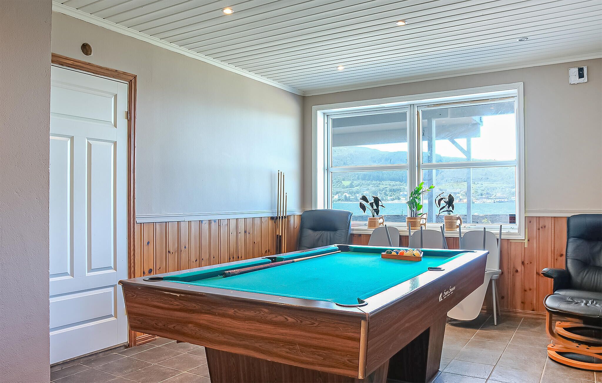 A pool table in a room with wooden paneling, a window with a view, and two chairs.
