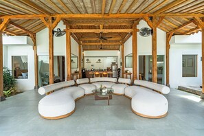 A covered outdoor seating area with a curved sofa, ceiling fans, and a kitchen in the background. - Brand New - 2bdrs: Luxury, Concierge & Security (Badung)