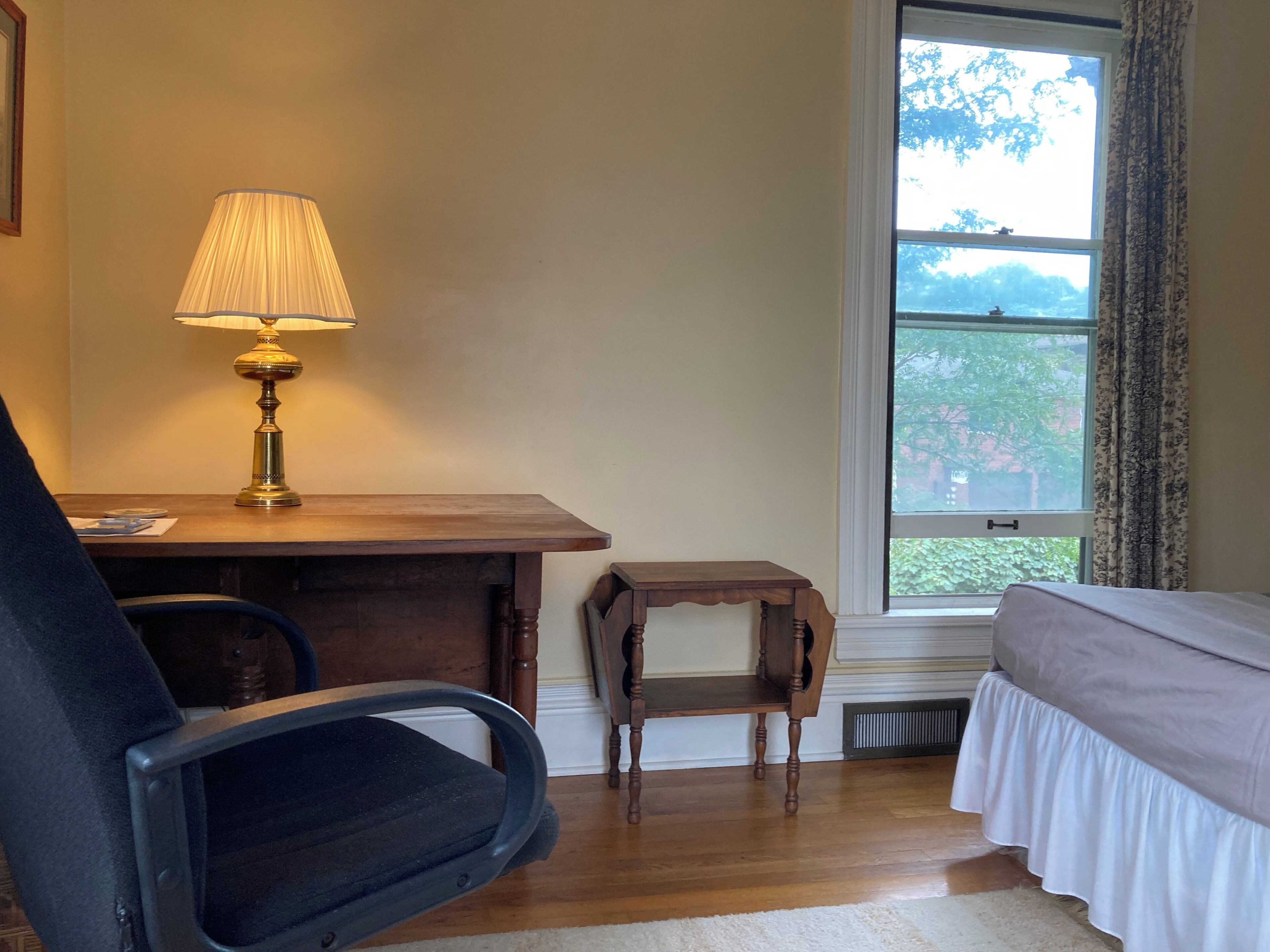 A room with a desk, chair, and a bed. There is a window with a view of trees.