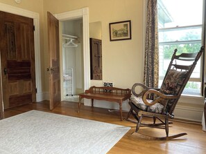 Ein Zimmer mit einem Holzschaukelstuhl, einem kleinen Tisch, einem weißen Teppich, einer Holztür, einem weißen Kleiderschrank und einem Fenster mit Vorhängen.