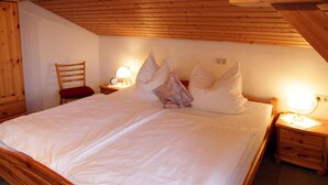 Uma cama com estrutura de madeira, roupa de cama branca, duas mesas de cabeceira com abajures e uma cadeira com almofada vermelha.