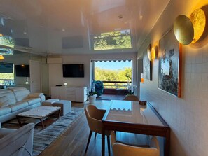 A modern living room with a sofa, coffee table, and dining table. There is a TV mounted on the wall, a balcony with a view of trees, and a kitchen area with cabinets and a countertop. - TOIT-TERRASSE D'EXCEPTION  et  VUE MER PANORAMIQUE A ST TROPEZ (ST TROPEZ)