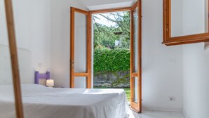Una camera da letto con un letto, un comodino, una lampada e una porta aperta su un giardino.