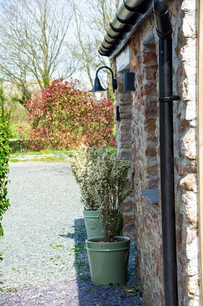 A stone building with a gravel path, potted plants, and a red flowering bush in the background.