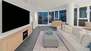 Una sala de estar moderna con una televisión de pantalla plana grande, un sofá, una mesa de centro y vistas de la ciudad a través de amplios ventanales.