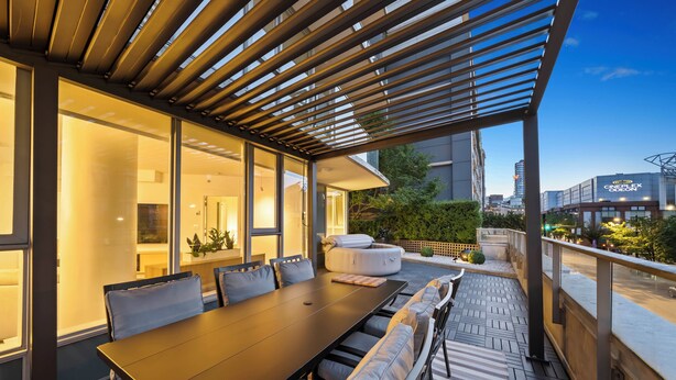 Un área de comedor al aire libre con una mesa larga, asientos acolchados y una pérgola arriba.