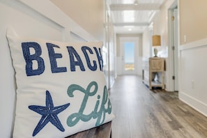 A hallway with a white wall, a wooden floor, and a pillow with 'BEACH Life' text. - Front Row Paradise | Pool & Ocean Views (Oak Island)