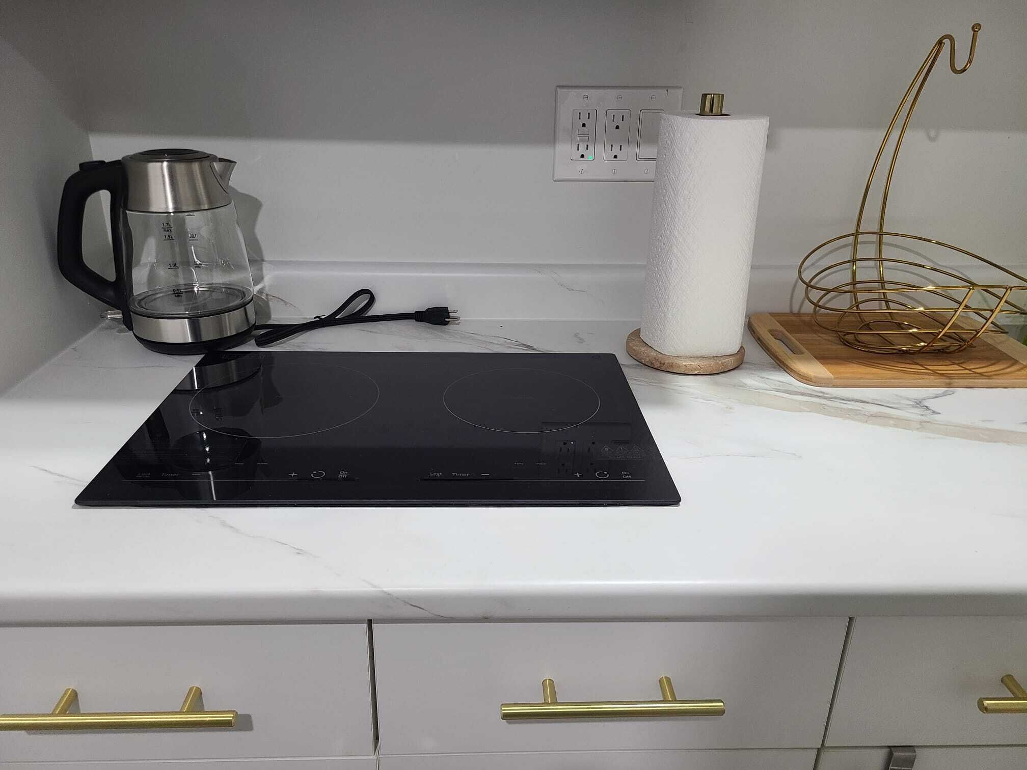 Cozinha com fogão por indução preto, chaleira de vidro, porta-papel toalha e cesto de arame dourado.