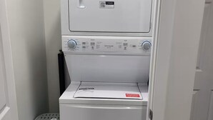 A compact washer and dryer unit in a small laundry room with a gray laundry basket and a mop head visible. - Haven Serene - Modern, convenient, quiet, private 1 bedroom basement suite (Winnipeg)