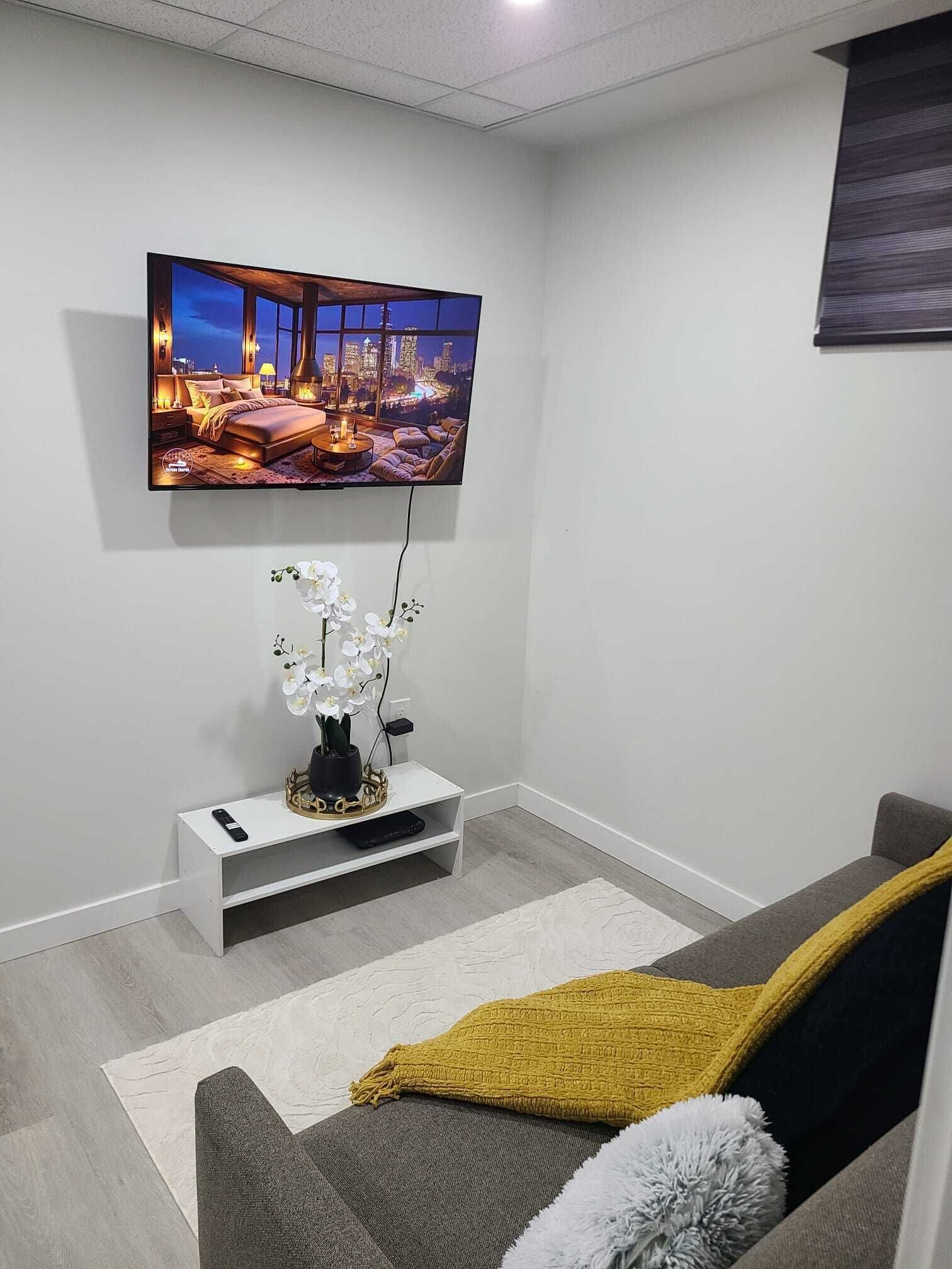 Uma sala de estar moderna com uma televisão de tela plana na parede, uma mesa de console branca com uma orquídea em vaso e um sofá cinza com um cobertor mostarda.
