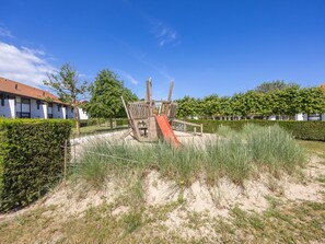Ein Spielplatz mit Rutsche und Klettergerüst, umgeben von Sand und Grünflächen, mit Gebäuden im Hintergrund.