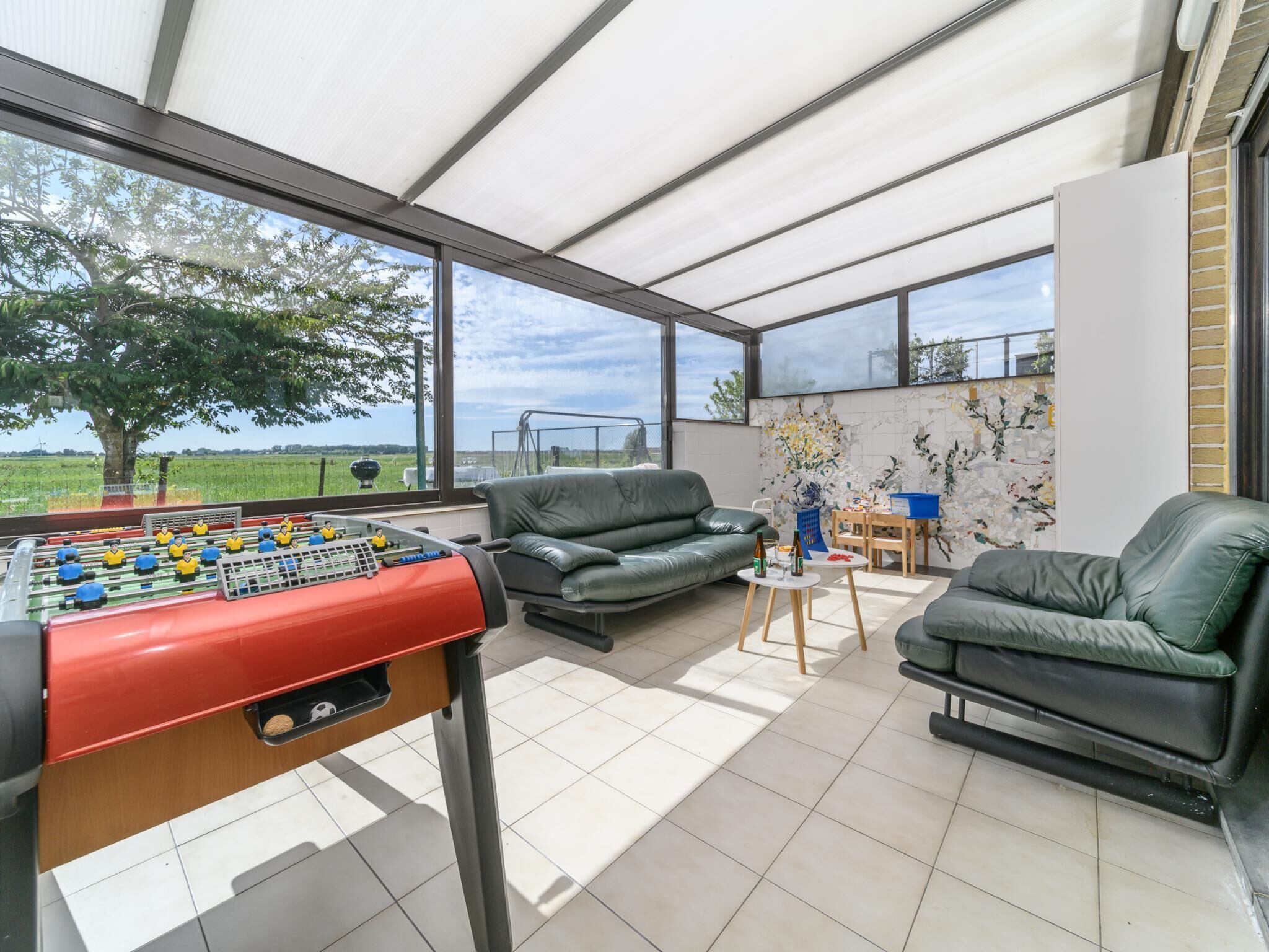 Une pièce spacieuse équipée d’un baby-foot, de deux canapés en cuir, d’une petite table avec des boissons, et offrant une vue sur un arbre et des champs à travers de grandes fenêtres.