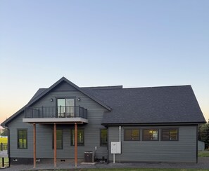 A modern two-story house with a balcony, a visible electrical box, and a gravel driveway.