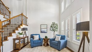 Een woonkamer met een houten trap, twee blauwe fauteuils, een salontafel, een vloerkleed en een schilderij aan de muur.
