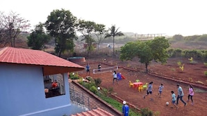 Uma área residencial com um edifício azul, uma construção com telhado vermelho e um terreno de terra onde pessoas estão brincando.