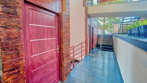 Una puerta roja con tiradores horizontales, pared de piedra, suelo azul y una escalera que lleva a un balcón con plantas en macetas.