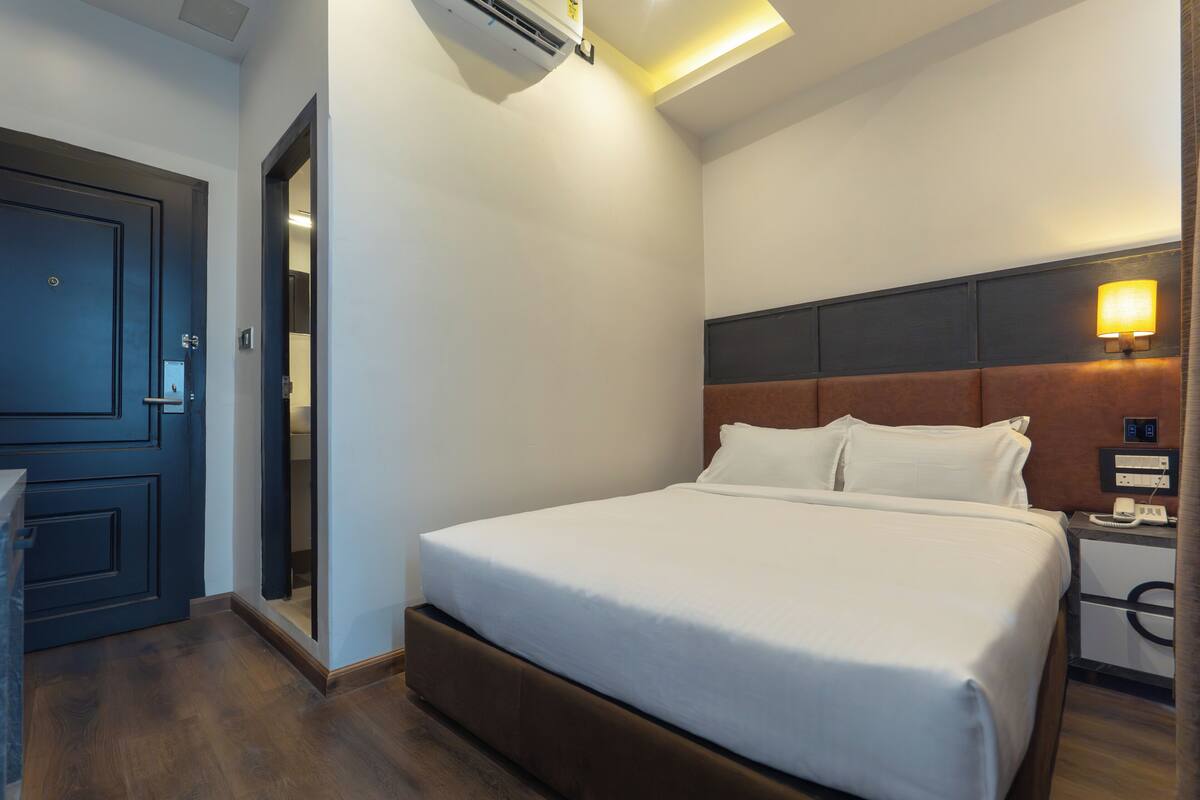 A hotel room with a bed, bedside table, lamp, and an air conditioning unit.