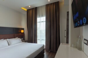 Quarto de hotel com cama, mesa de cabeceira, televisão e janela com cortinas.