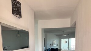 A hallway with a wooden floor, a white wall with a decorative panel, a mirror, a small shelf with decorative items, and a blue sofa in the background. - Beach House. Beachfront Apartment (Creixell)