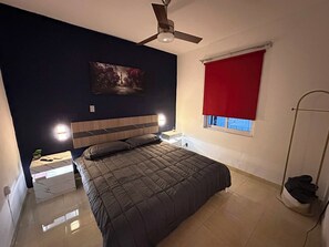 Ein Schlafzimmer mit einem Bett, einem Deckenventilator, einem Nachttisch, einem roten Fensterrollo und einem an der Wand hängenden Bild.