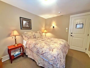 A bedroom with a bed, two bedside lamps, a framed picture on the wall, a door with a notice, and a small wooden nightstand with a lamp.