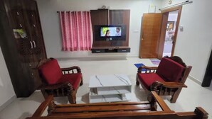 Ein Wohnzimmer mit Fernseher, zwei Holzstühle mit roten Kissen, einem weißen Couchtisch und einem Deckenventilator.