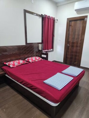 A bed with a red bedspread, two pillows, a wooden headboard, a mirror, a red curtain, a wooden door, and an air conditioning unit.