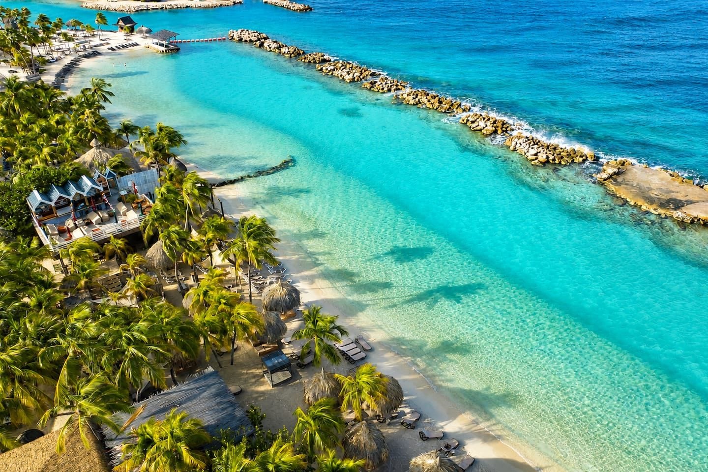 A resort with a clear turquoise sea, palm trees, and a sandy beach.