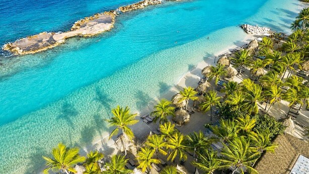 Vista aérea de una playa con aguas turquesas cristalinas, palmeras y tumbonas.