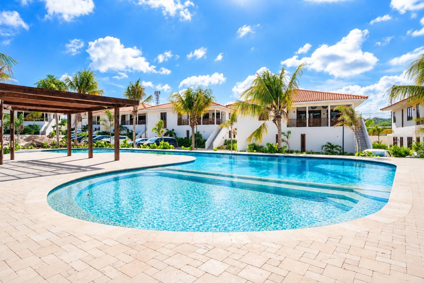 A swimming pool with a curved edge, surrounded by a paved area with a pergola structure.