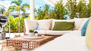 Un sofá blanco con cojines, una mesa redonda de madera con dos plantas en macetas y una mesita de mimbre con una lámpara.
