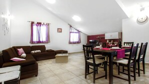 Una sala de estar con sofá, una mesa de comedor con sillas, una zona de cocina y una ventana con cortinas.
