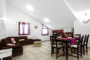 Ein Wohnzimmer mit Sofa, einem Esstisch mit Stühlen, einer Küchenzeile und einem Fenster mit Vorhängen.