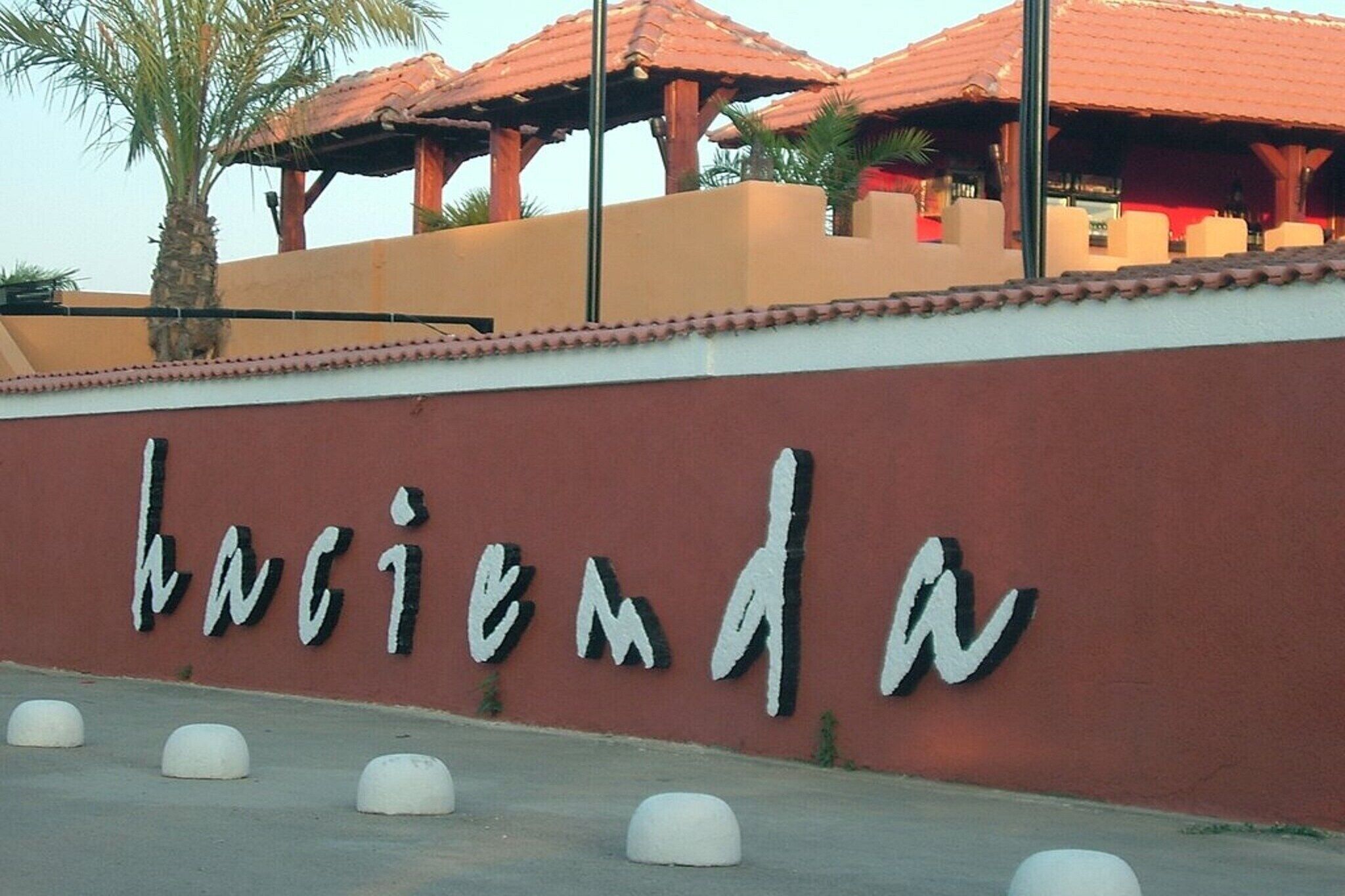 Ein Schild mit dem Wort 'hacienda' an einer Wand, Palmen und ein Gebäude mit rotem Ziegeldach.