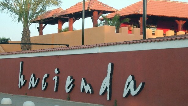 Un letrero con la palabra 'hacienda' en una pared, palmeras y un edificio con tejado de tejas rojas.