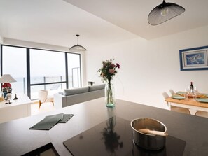 A modern kitchen with a large island, a dining area with a wooden table, and a view through large windows. - Luxurious Flat With Magnificent sea View (Porspoder)